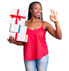 Wall Mural - Young african american woman holding gift doing ok sign with fingers, smiling friendly gesturing excellent symbol