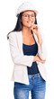 © Krakenimages.com - Young beautiful latin girl wearing architect hardhat and glasses looking confident at the camera with smile with crossed arms and hand raised on chin. thinking positive.