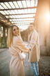 A young couple in beige trench coats walks through the park and has fun. A woman and a man pose on an alley on a sunny spring day at sunset.
