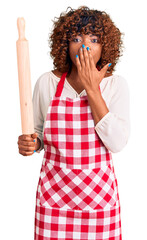 Sticker - Young african american woman wearing apron holding rolling pin covering mouth with hand, shocked and afraid for mistake. surprised expression