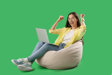 Young female programmer with laptop celebrating success while sitting on beanbag chair against green background