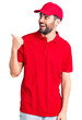 © Krakenimages.com - Young handsome man with beard wearing delivery uniform smiling with happy face looking and pointing to the side with thumb up.
