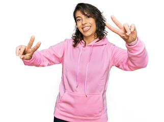 Wall Mural - Young hispanic woman wearing casual sweatshirt smiling with tongue out showing fingers of both hands doing victory sign. number two.
