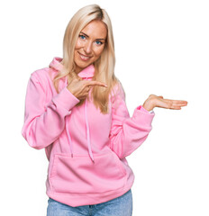Wall Mural - Young blonde woman wearing casual sweatshirt amazed and smiling to the camera while presenting with hand and pointing with finger.