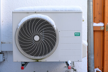 Outdoor heat pump unit covered with snow and R32 refrigerant label sitting on sound and vibration insulating rubber pads against a white stucco house exterior next to a wooden fence and zinc rain pipe