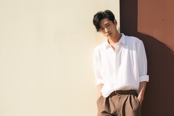 Poster - A handsome young Asian man in a white shirt and brown pants is standing against a wall.