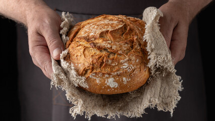 Wall Mural - Man baker holding fresh spelt wheat loaf of bread on rustic linen towel on dark background. Healthy pastry.