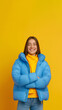 © Zoran Karapancev - Smiling happy brunette teenage girl wears blue puffer jacket and yellow turtleneck, crossed hands, blue jeans, isolated on yellow backdrop, copy space, vertical 9:16