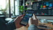© kitidach - Investor checking stock market trends on a smartphone with financial data graphs, in an office with multiple computer screens.