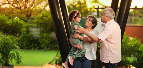 Indian Asian happy aged old elder male man woman young female lady standing hold cute child baby girl kid play outdoor home park older lover couple family funny talk enjoy fun joy outside house garden