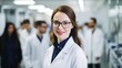 © Ameer - Portrait of a confident female researcher in a white lab coat and glasses, working in a modern medical science laboratory with a team of specialists in the background