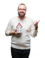 Sticker - Young caucasian real state agent man holding house isolated background pointing and showing with thumb up to the side with happy face smiling