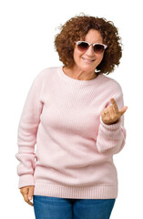 Sticker - Beautiful middle ager senior woman wearing pink sweater and sunglasses over isolated background Beckoning come here gesture with hand inviting happy and smiling