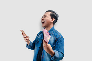 Young Asian man holding money and this phone showing happy expression when looking to empty space