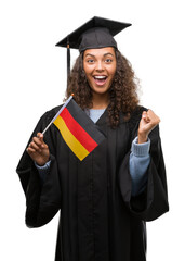 Wall Mural - Young hispanic woman wearing graduation uniform holding flag of Germany screaming proud and celebrating victory and success very excited, cheering emotion