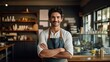 © AdriFerrer - A cheerful male barista with curly hair stands with crossed arms in a cozy cafe.
