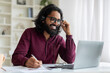© Prostock-studio - Young indian man wearing headset using laptop and smiling while taking notes