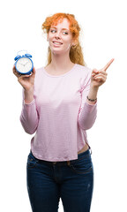 Poster - Young redhead woman holding alarm clock very happy pointing with hand and finger to the side