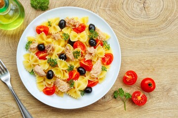 Farfalle pasta salad with canned tuna in olive oil, cherry tomatoes,black olives,parleys,olive oil and peppers on plate with wooden background.Healthy Italian summer salad.Top view.Copy space

