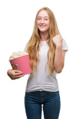 Poster - Blonde teenager woman eating pop corn screaming proud and celebrating victory and success very excited, cheering emotion