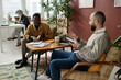© AnnaStills - Biracial male coworkers discussing documents with infographics sitting at table in office