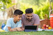 Happy smiling indian father with son using digital tablet during weekend picnic at park - concept of cyberspace, family fun and Joyful interaction