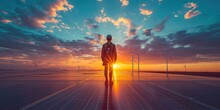 Male Green Energy Engineer Walking On Solar Panel, Wearing Safety Belt And Hard Hat, Inspecting Sustainable Energy Farm With Wind Turbines, Sunset, Flare Light