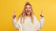 © Lila Patel - Young Blonde Caucasian Woman Wearing a White Sweatshirt Poses on a Yellow Studio Background, Expressing Excitement by Pointing Her Fingers, Radiating Positive and Dynamic Energy