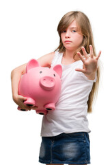 Sticker - Young blonde toddler holding piggy bank with open hand doing stop sign with serious and confident expression, defense gesture