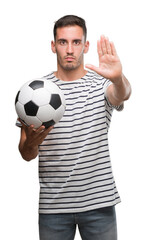 Poster - Handsome young man holding soccer football with open hand doing stop sign with serious and confident expression, defense gesture