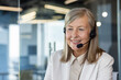 © Liubomir - Close-up photo of a senior business woman sitting in the office and smiling while working wearing a headset.