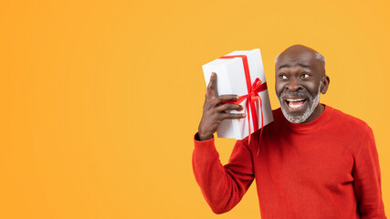 Wall Mural - Joyful elderly African American man in a red sweater excitedly holding a white gift box