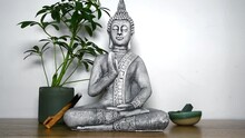 High Definition Video In White Background Of A Buddha Figurine In Meditative Position, On One Side, There Are Decorative Elements Such As A Plant And A Green Stone On A Pot. 