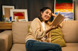 © Chanakon - Asian beautiful woman sitting reading book on cozey couch sofa in living room with warm light at night. People female stay home part time relax at home.