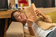© Chanakon - Asian beautiful woman lay down on cozey couch sofa reading book in living room with warm light at night. People female stay home part time relax at home.