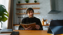 Young Man Student With Beanie Sitting In Modern Kitchen At Home Looking At The Notes In Notebook He Is Holding In His Hand, Reading, Thinking And Changing The Pages	
