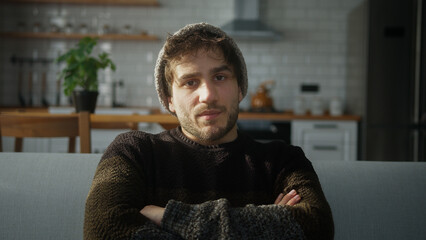 Bored young man with beanie sitting on sofa at home with kitchen background, having a conference call, listening to the speaker on the video call
