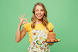 Young housewife housekeeper chef cook baker woman wear apron yellow t-shirt hold in hand plate with pancakes show ok okay wink isolated on plain pastel green background studio. Cooking food concept.
