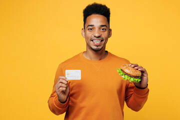 Wall Mural - Young happy man wear orange sweatshirt casual clothes eat burger hold credit bank card isolated on plain yellow background studio portrait. Proper nutrition healthy fast food unhealthy choice concept.