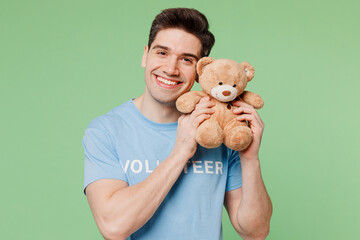 Sticker - Young happy smiling man he wears blue t-shirt white title volunteer hold hug teddy bear plush toy isolated on plain pastel green background. Voluntary free work assistance help charity grace concept.