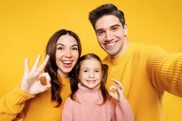 Wall Mural - Close up young parents mom dad with child kid girl 7-8 years old wear pink sweater casual clothes do selfie shot mobile cell phone show ok okay isolated on plain yellow background. Family day concept.