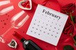 Love is near: Top view of February calendar, chocolate treats, wine bottle, glasses, roses, silk ribbon, and confetti on a passionate red background. Create your Valentine's message