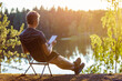 A man is reading an e-book on the shore of a forest lake at sunset in the evening.
