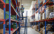 © Nassorn - Warehouse worker standing on ladder checking stock on shelf. Female blue collar staff in safety uniform standing on staircase working with merchandise on storage rack in store. Delivering service job.
