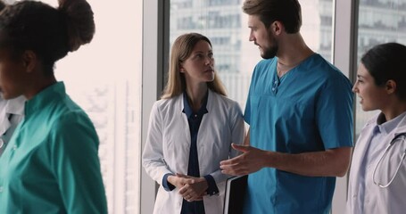 Wall Mural - Team of serious diverse medical colleagues in white and blue uniforms walking together past camera in hospital office hall, talking, discussing difficult job cases, career in medicine, teamwork