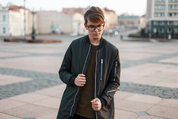 Wall Mural - Stylish urban handsome guy with glasses with a hairstyle in a fashion black jacket walks in the city