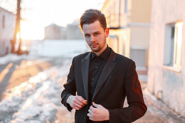 handsome successful businessman guy with a hairstyle in a fashion black suit wears a blazer and stands on the street at sunset