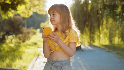 Wall Mural - Preteen child kid using smartphone typing text answering messages chatting online looking mobile screen social media app, playing game. Pretty blonde girl standing on city sunset park street, outdoors