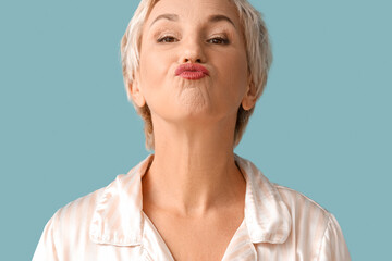 Wall Mural - Beautiful mature woman in pajamas doing face building exercise on blue background, closeup