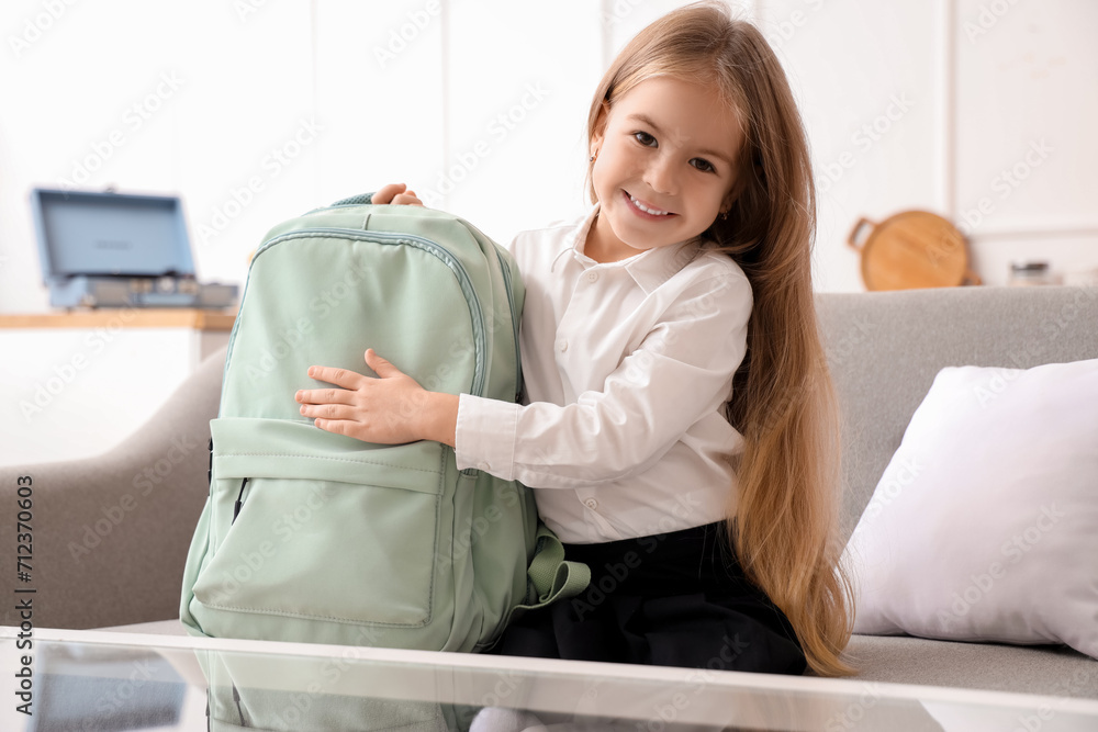 Cute little schoolgirl with backpack sitting on sofa at home